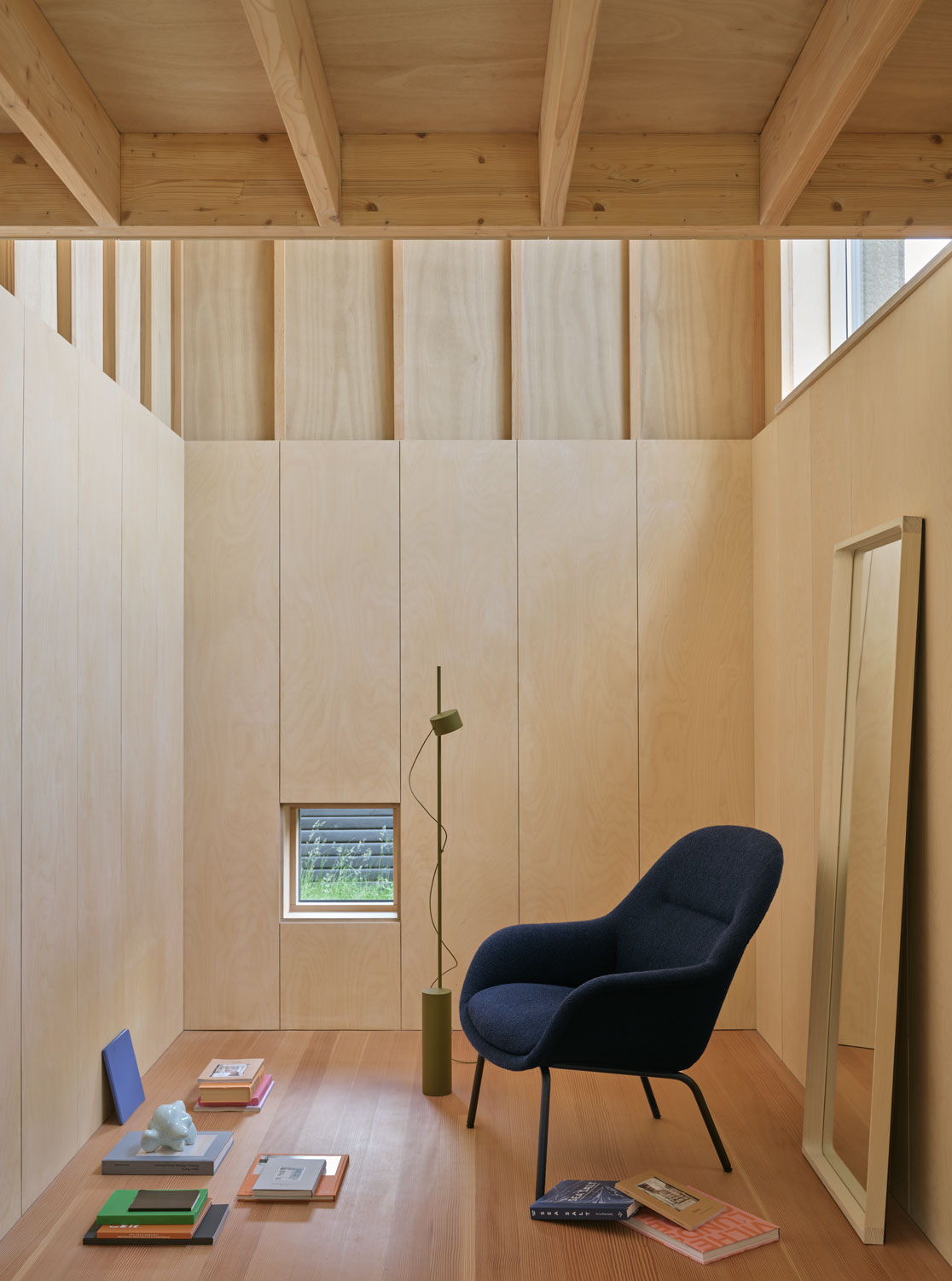 A modern Muuto interior featuring an Arced Mirror with a spruce frame, paired with a Fiber Lounge Chair with a tube base. The setup includes a brown green Post floor lamp, adding a warm, ambient light to the space. The combination of elements creates a sophisticated and contemporary look.