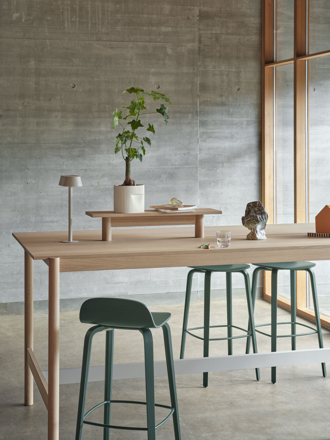 A Muuto setup featuring a Linear System high table 105 cm and tray 75 cm in oak, paired with a Visu bar stool 75 cm in dark green. The scene includes a Ridge planter 15 cm in off-white and a Linear table lamp. The combination of modern furnishings and understated accessories creates a sophisticated and stylish space.