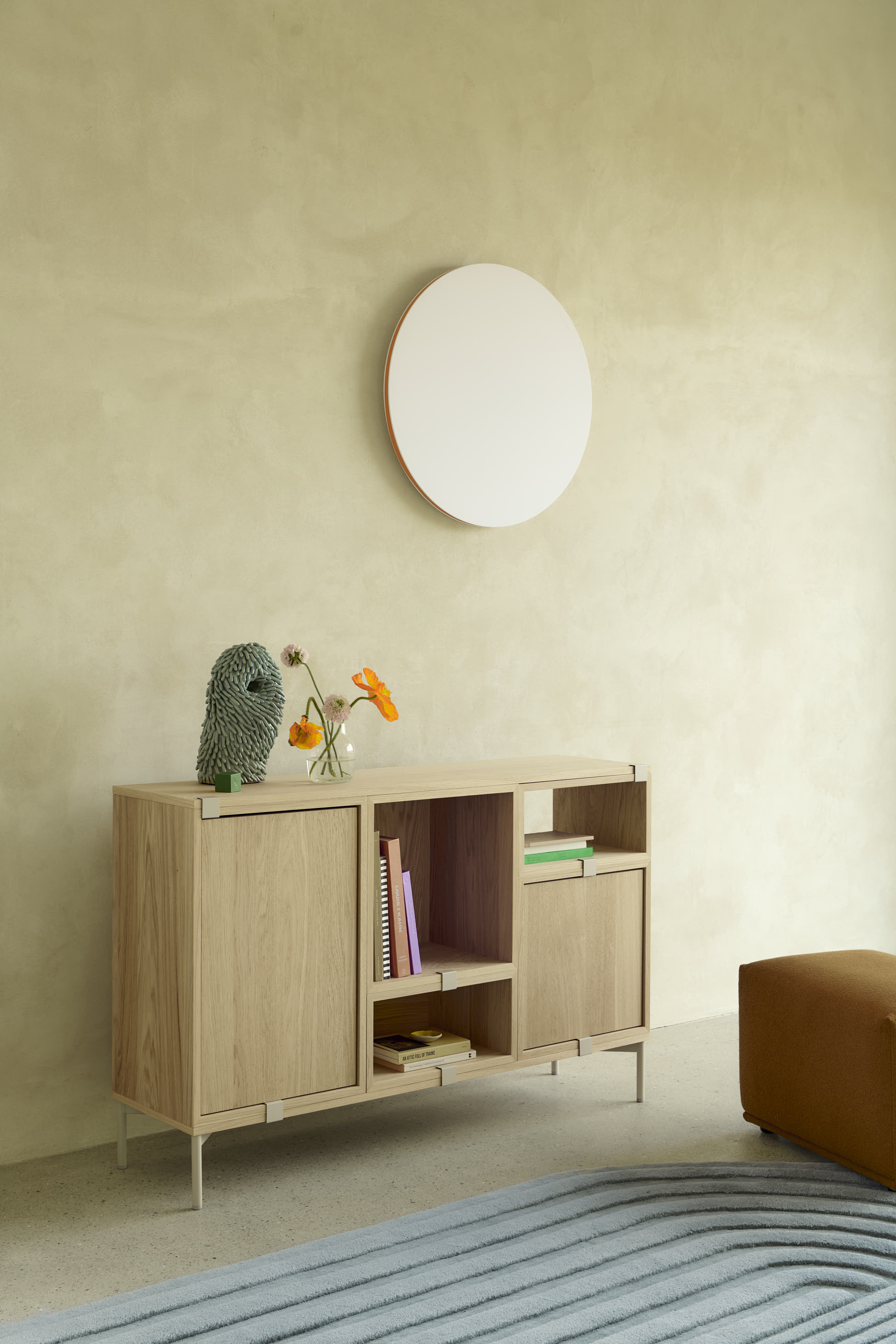 Stacked in oak is placed in a minimalistic room with brushed stone walls. A round wall lamp hangs above the storage system, casting a soft light that adds a subtle, elegant touch to the clean design.