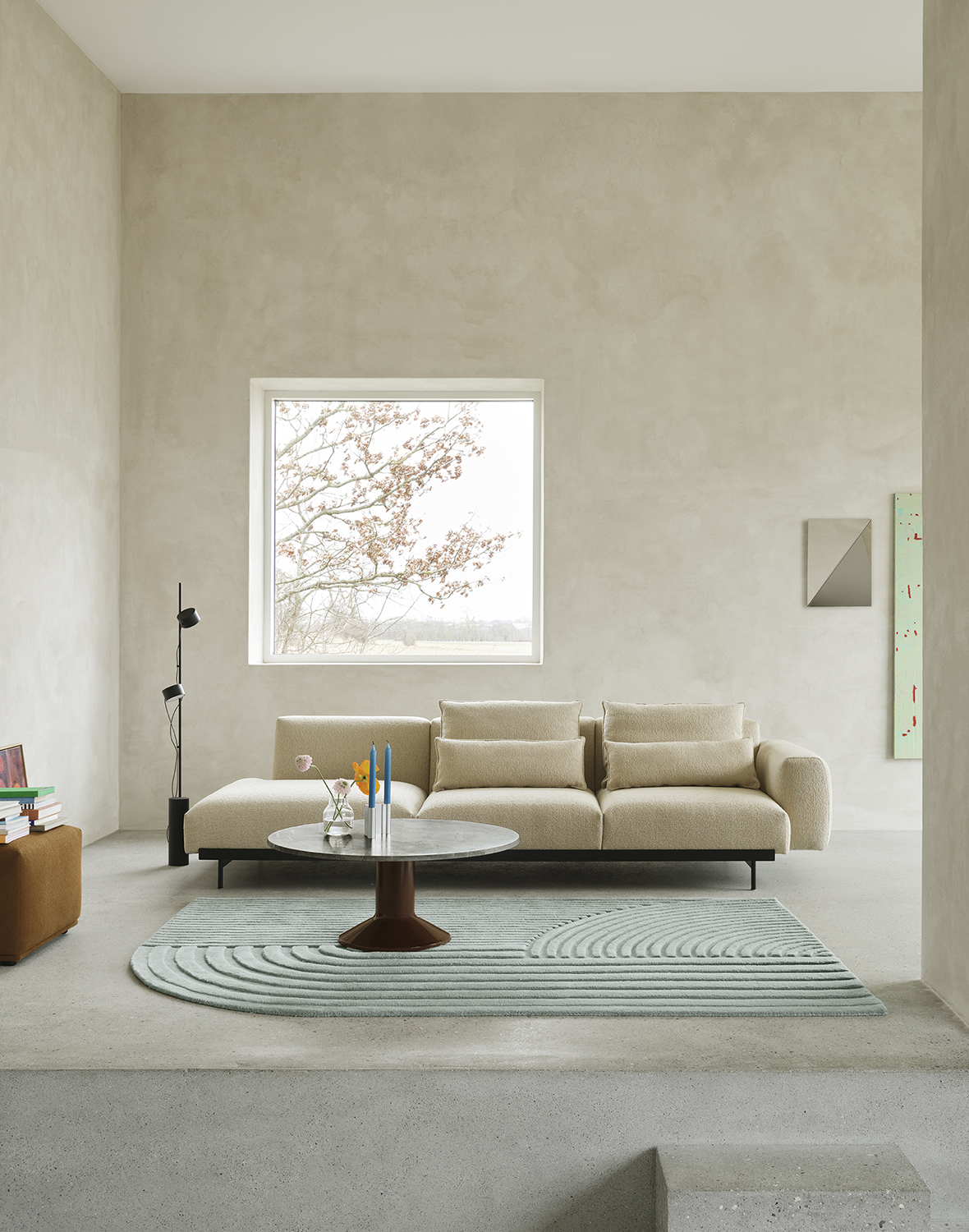 A sand-colored sofa is positioned against a brushed grey stone wall. A black floor lamp stands beside the sofa, while a round coffee table in marble and burgundy is placed in front, atop a light blue rug. The arrangement creates a stylish and serene living space.