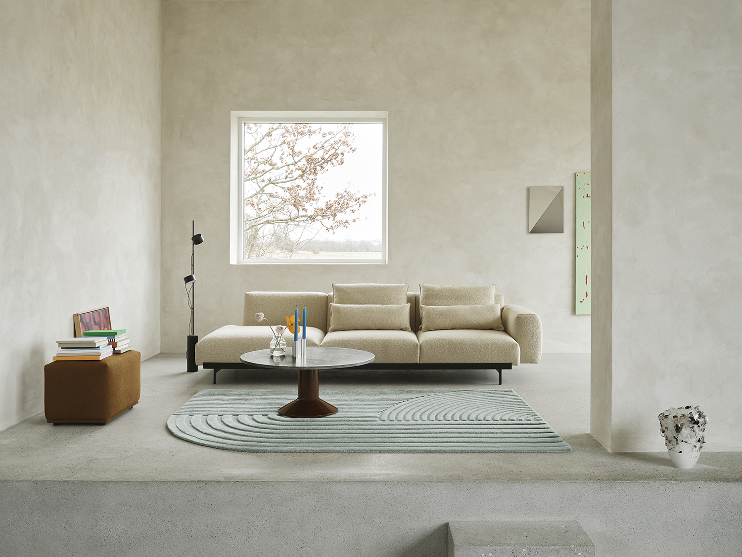 A sand-colored sofa is positioned against a brushed grey stone wall. A black floor lamp stands beside the sofa, while a round coffee table in marble and burgundy is placed in front, atop a light blue rug. The arrangement creates a stylish and serene living space.