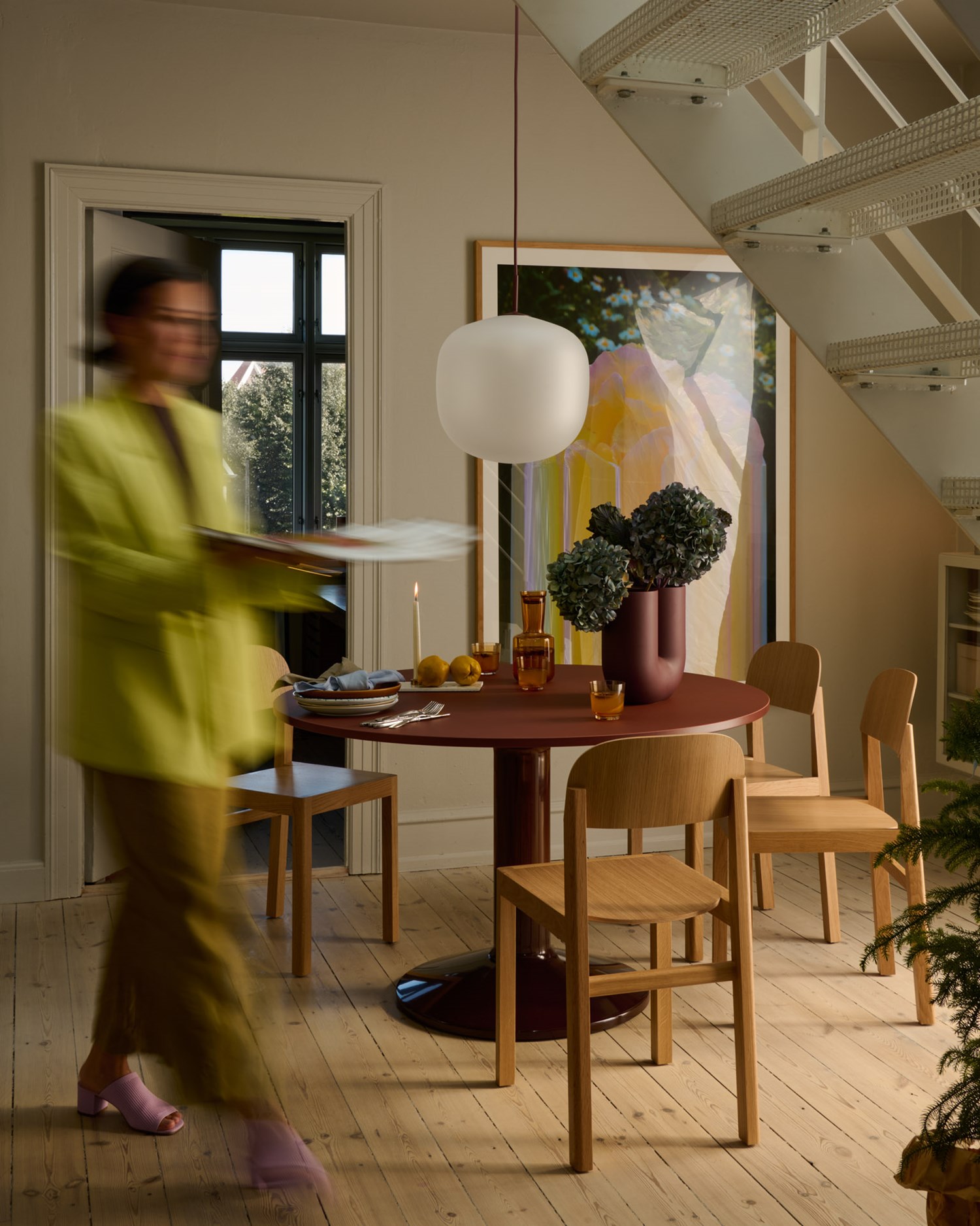 A woman walks past a round deep red table holding a vase with kale, two glasses, and plates. A soft light from a large window highlights the warm and cozy atmosphere, with a large artwork visible in the background. A woman walks past a round deep red table holding a vase with kale, two glasses, and plates. A soft light from a large window highlights the warm and cozy atmosphere, with a large artwork visible in the background.